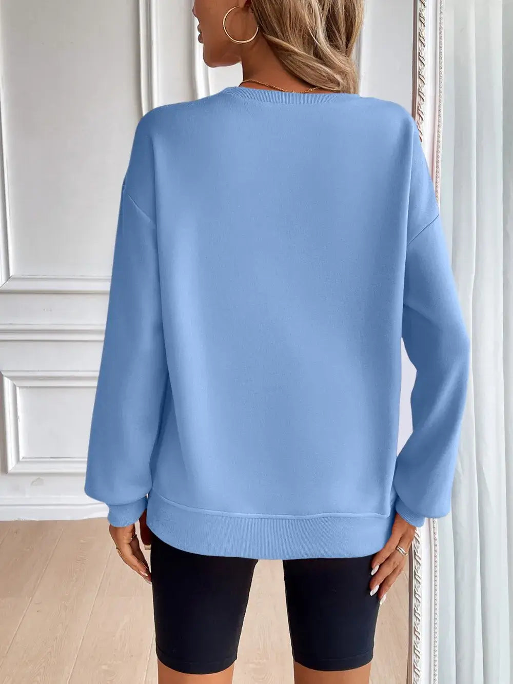 Back view of a woman wearing a light blue Ivy Lane round neck long sleeve sweatshirt paired with black shorts.