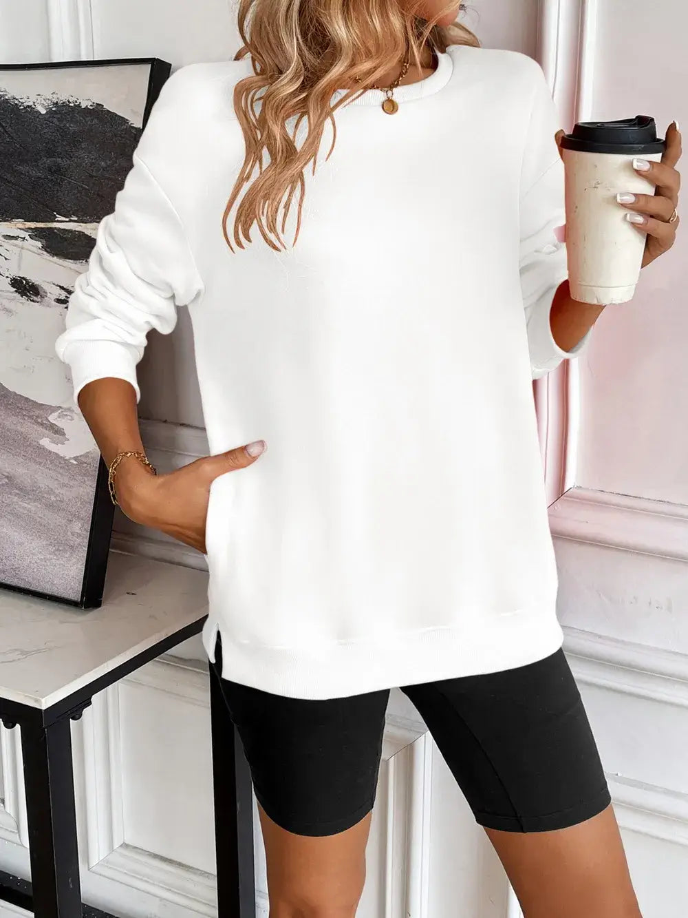 Woman wearing white Ivy Lane round neck long sleeve sweatshirt with black shorts, holding a coffee cup.