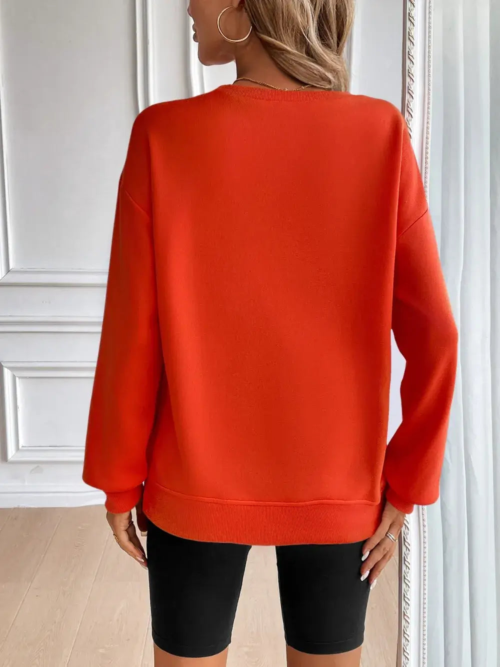 Back view of a woman wearing an orange Ivy Lane round neck long sleeve sweatshirt, paired with black shorts.